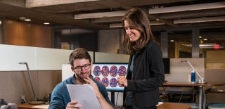 Professor Emily Falk talking to a student, looking at a paper. Behind them there are brain scans on a monitor.