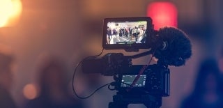 Focused shot of a videographers camera with an attached mic. On the display screen, there is what seems to be a wedding reception going on with guests standing in a circle. Around the camera is completely blurred. Photo credit for Kushagra Kevat, Unsplash.