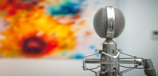 Side view of a silver microphone in focus. In the blurred background is an artwork with shades of the colors yellow, red, and blue. Photo credit for Michal Czyz,Unsplash.