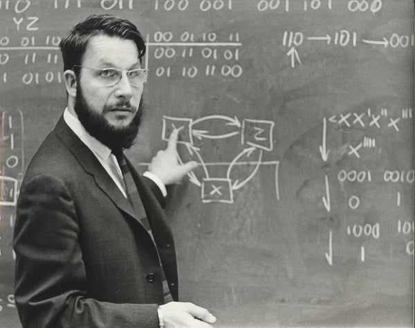 Man in a suit pointing at a blackboard that has various mathematical drawings