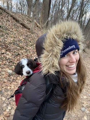 Person in Annenberg hat and winter coat in the woods, carrying a dog in a backpack